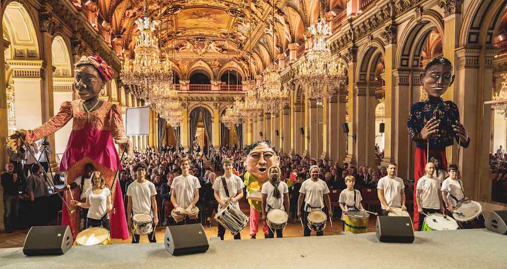 Giant Mozambique puppets, together with the band from Parade, at an earlier presentation in March 2019, all participants will march from Hôtel de Ville to the theatre on Friday next ©Thomas Amouroux, Théâtre du Châtelet
